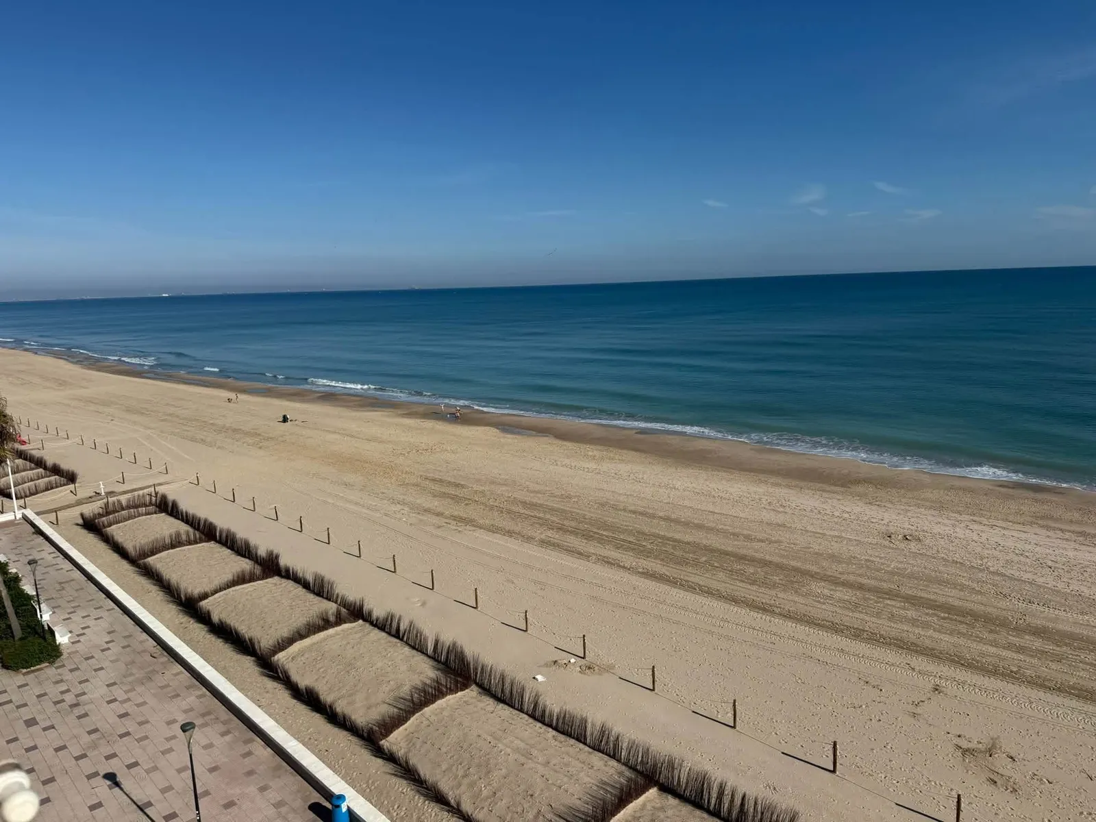 Vista desde el apartamento de la amplia playa de la Llastra, las dunas protegidas junto al paseo y el mar en calma bajo un cielo azul